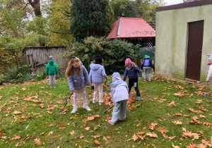 Na trawniku w ogrodzie dzieci zgrabiają kolorowymi grabkami jesienne liście w kupki. W tle widać drewniany płot, altanę z czerwonym dachem, krzewy i budynek z brązowymi drzwiami.
