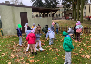 Dzieci w kurtkach i czapkach grabią i zbierają opadłe dębowe liście na trawie w ogrodzie przedszkolnym, przy niskim budynku z zadaszeniem i drewnianym płotkiem. W tle widać plac zabaw z huśtawką, drewnianą lokomotywą i budynek przedszkola.