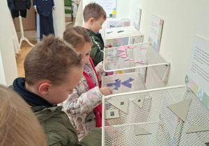 Dzieci układają kolorowe trójkątne elementy przypominające tangram na białych siatkowych półkach zamocowanych do ściany w sali muzealnej z tablicą „Instrukcja”. W tle stoją manekiny w strojach z dawnej epoki: ciemny frak, damska suknia z kapeluszem i biała suknia.
