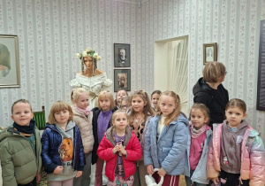 Grupa dzieci z opiekunką stoi w sali muzealnej z tapetą w kwiatowe paski, obok manekina w jasnej sukni z dawnej epoki i wianku z kwiatów. Na ścianach wiszą oprawione portrety i fotografie, a na podłodze leży wzorzysty dywan.