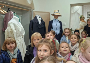 Grupa dzieci zwiedza wystawę historycznych strojów w muzeum; w tle widać manekiny w sukniach, fraku i kostiumie z kapeluszem oraz wieszak z ubraniami.