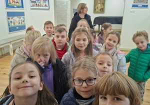 Dzieci w kurtkach stoją w grupie wraz z opiekunką w jasnej sali wystawowej, na której ścianach wiszą kolorowe prace w stylu ludowym oraz portret w złotej ramie. W głębi sali stoi czarny fortepian.
