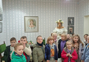 Grupa dzieci w kurtkach pozuje do wspólnego zdjęcia w muzealnej sali stylizowanej na XIX-wieczny salon, z tapetą w kwiatowe pasy, zielonymi meblami i dywanem. Za nimi stoi manekin w białej sukni z wiankiem z kwiatów, a na ścianach wiszą portrety w ramach.