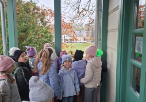 Grupa dzieci w czapkach i kurtkach stoi na zadaszonym, drewnianym ganku z zielonymi słupkami, obok zielonych, przeszklonych drzwi. Za nimi widać jesienne drzewa, trawnik i budynki w oddali.