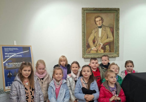 Grupa dzieci w kurtkach pozuje do wspólnego zdjęcia w sali muzealnej, pod oprawionym w złotą ramę portretem mężczyzny w XIX-wiecznym stroju, obok przykrytego czarnym pokrowcem fortepianu. Po lewej stoi plakat zapraszający na „Wystawę pokonkursową III Triennale Wycinanki Ludowej Województwa Łódzkiego”.