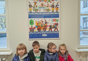 Czworo dzieci w kurtkach siedzi na ławce w jasnej sali galerii, pod oprawionym kolorowym wycinankowym obrazem w stylu łowickim. Na obrazie są postacie w strojach ludowych przy stole, kwiaty oraz wóz zaprzężony w konia.