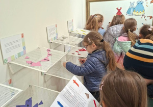 Dzieci oglądają w muzeum ścienną ekspozycję edukacyjną: na siatkowych półkach leżą układanki tangram z kolorowych kształtów, a nad nimi wiszą tabliczki z opisami. Jedno dziecko układa elementy, inne trzyma ilustrowaną broszurę z kolorowymi figurami, a w tle widać tablicę z papierowymi sukienkami do przypinania.