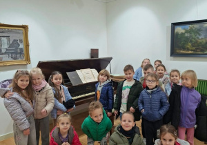 Grupa dzieci pozuje do wspólnego zdjęcia w sali muzealnej przy zabytkowym fortepianie z otwartymi nutami na pulpicie. Na ścianach wiszą obrazy w złoconych ramach, w tym pejzaż z drzewami i obraz przedstawiający fortepian.
