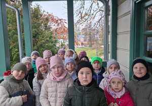 Grupa przedszkolaków w czapkach i kurtkach stoi na zadaszonej werandzie drewnianego budynku z zielonymi słupami. W tle widać jesienne drzewa i ceglany dom.
