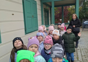 Grupa dzieci w czapkach i kurtkach stoi w rzędzie przed jasnym drewnianym budynkiem z zielonymi okiennicami i zadaszonym gankiem, a obok idzie opiekunka. Zdjęcie zrobiono na wybrukowanej ścieżce podczas pochmurnej wycieczki.
