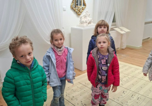 Grupa dzieci stoi w jasnej sali muzealnej z białymi draperiami, złotym lustrem na ścianie i białą rzeźbą na postumencie. Podłogę pokrywa duży wydruk rękopisu nutowego z odręcznymi napisami, m.in. „Oboe” i „Clarinetti”.