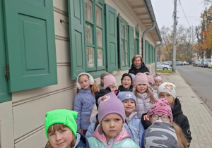 Grupa dzieci w czapkach i kurtkach stoi w rzędzie przed jasnym drewnianym budynkiem z zielonymi okiennicami i zadaszonym gankiem, a obok idzie opiekunka. Zdjęcie zrobiono na wybrukowanej ścieżce podczas pochmurnej wycieczki.