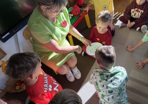 Nauczycielka pokazuje dzieciom pestki jabłek na białym talerzyku.