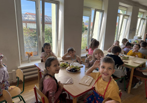 Grupa dzieci w jasnej sali przedszkolnej lub szkolnej siedzi przy drewnianych stołach z deskami do krojenia i talerzami owoców (winogrona, śliwki, jabłka), trzymając drewniane nożyki.