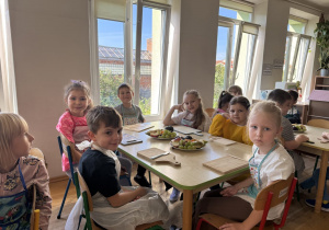 Grupa dzieci w fartuszkach siedzi przy stołach w jasnej sali przedszkolnej, przy dużych oknach. Na stołach znajdują się talerze z pokrojonymi owocami (jabłka, winogrona, śliwki), drewniane deski do krojenia i noże.
