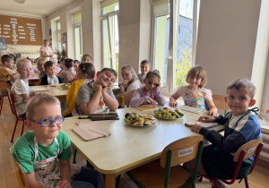 Grupa dzieci w fartuszkach siedzi przy stołach w sali przedszkolnej, przed nimi leżą drewniane deski do krojenia oraz talerze z winogronami i pokrojonymi owocami.