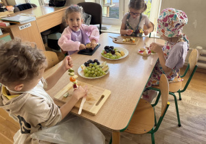 Dzieci w fartuszkach nadziewają kawałki owoców (winogrona, śliwki, jabłka, gruszki) na patyczki przy stole w sali, korzystając z drewnianych deseczek do krojenia. Na stole stoją talerze z owocami, a w tle widać biurko z tabliczką „BIURKO”.
