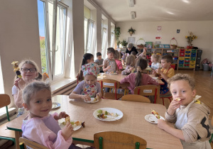 Grupa dzieci w kolorowych fartuszkach siedzi przy drewnianych stolikach w sali przedszkolnej, jedząc szaszłyki owocowe z talerzyków.