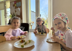 Troje dzieci w kucharskich fartuszkach siedzi przy stole i trzyma szaszłyki z owocami (m.in. winogrona, jabłko), a przed nimi stoją białe talerzyki.