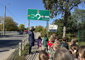 Grupa dzieci z opiekunką stoi na chodniku przy ulicy i ogląda drogowskaz z napisami „Jeżów”, „Łódź”, „Centrum” i „Łyszkowice” oraz symbolem ronda. W tle widać jezdnię z samochodami, drzewa i błękitne niebo.