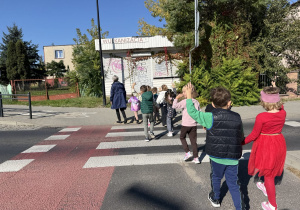 Grupa dzieci wraz z opiekunką przechodzi przez oznaczone pasy dla pieszych na ulicy. W tle widoczny jest zamknięty budynek z szyldem „WULKANIZACJA” oraz drzewa i krzewy.