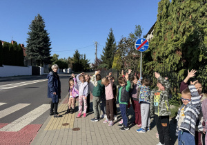 Grupa dzieci z opiekunką stoi na chodniku przy przejściu dla pieszych, obok znaku drogowego oznaczającego koniec drogi dla pieszych i rowerów. Zajęcia wyglądają na praktyczną lekcję bezpieczeństwa w ruchu drogowym.