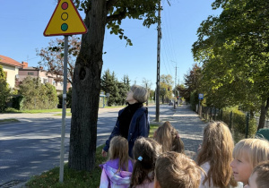 Grupa dzieci z opiekunką stoi na chodniku przy ulicy i ogląda żółto-czerwony trójkątny znak drogowy ostrzegający o sygnalizacji świetlnej, umieszczony pod drzewem.