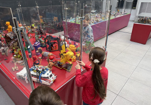Dzieci oglądają wystawę modeli z klocków w przeszklonych gablotach na czerwonych stołach, m.in. figurkę Pikachu, Eevee, Pokeball, samochód Ecto-1 z Ghostbusters oraz miniaturowe miasteczko. Jedno z dzieci wskazuje palcem na eksponaty za szybą.