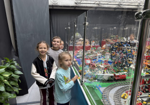 Dzieci oglądają dużą makietę z klocków LEGO za plastikową barierką, z miasteczkiem, diabelskim młynem, torami kolejowymi i pociągiem. Jedno z dzieci trzyma żółty element, a w tle widać kolejne gabloty z budowlami.