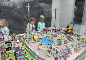 Dzieci oglądają przez plastikową gablotę dużą makietę z klocków LEGO Friends: kolorowe domki, basen z delfinami, statek, kawiarnię i jeżdżący po torach pociąg. Wystawa znajduje się we wnętrzu budynku.
