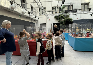 Grupa dzieci wraz z opiekunką ogląda wystawę budowli z klocków LEGO umieszczonych w przeszklonych gablotach na czerwonych i niebieskich stołach w przestronnej, oszklonej hali ze ścianą zieleni. Wśród eksponatów widać kolorowe miniaturowe miasta i statki kosmiczne.