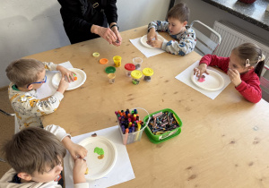 Dzieci siedzą przy drewnianym stole w sali i wykonują pracę plastyczną: na papierowych talerzykach układają kolorowe elementy z ciastoliny według narysowanych konturów. Na stole stoją pojemniki z ciastoliną, kubek z flamastrami i pudełko z kredkami świecowymi, a osoba dorosła pomaga przy stole.