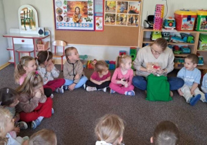 Dzieci siedzą w kole na dywanie w sali przedszkolnej i słuchają nauczycielki, która wyjmuje przedmiot z zielonego worka. W tle widać półki z zabawkami, pojemniki z klockami i tablicę korkową z plakatem „Jesienny krajobraz”.