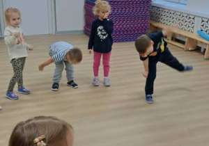 Grupa dzieci ćwiczy w jasnej sali z drewnianą podłogą: kilkoro z nich robi skłony i wymachy nóg, a jedno stoi na pierwszym planie i klaszcze. Pod ścianą leży wysoki stos kolorowych mat gimnastycznych, a pod oknem stoi drewniana ławka.