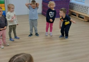 Grupa dzieci stoi w półokręgu w jasnej sali z drewnianą podłogą i wykonuje ćwiczenia ruchowe, na przykład trzyma ręce na głowie albo pochyla się do przodu. W tle widać stos kolorowych mat gimnastycznych, drewnianą ławkę pod oknem i przeszklone drzwi.