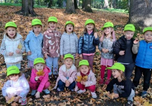 Grupa dzieci w zielonych czapeczkach pozuje do wspólnego zdjęcia w jesiennym parku, wśród drzew i opadłych liści. Część dzieci trzyma w rękach żółte liście klonu.