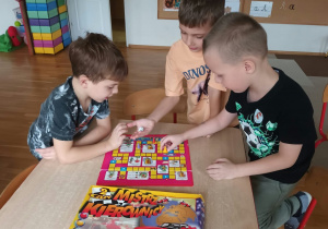 Troje dzieci gra przy stoliku w sali przedszkolnej w planszową grę „Mistrz Kierownicy”, rzucając kostką i przesuwając pionki po kolorowej planszy. Obok planszy leży pudełko od gry, a w tle widać krzesełka, kolorowe pojemniki i tablice z literami.