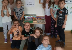Grupa dzieci pozuje w sali przedszkolnej przy stoliku z pudełkami gier planszowych, obok kartki z napisem „Kącik gier planszowych”. Na ścianie wiszą plakat „Kontrakt dobrego zachowania” ozdobiony kolorowymi odciskami dłoni oraz tablica dyżurów.