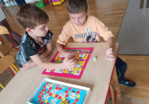 Dwoje dzieci gra przy stoliku w sali przedszkolnej w planszową grę z kostką i pionkami, przesuwając pionek po kolorowej planszy. Obok na stoliku leży otwarte pudełko z drugą grą planszową.