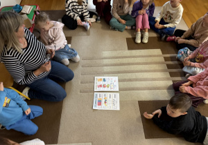 Zajęcia w przedszkolnej sali: dzieci i nauczycielka siedzą w kręgu na beżowo-brązowym dywanie. Na środku leżą dwie karty pracy z kolorowymi obrazkami owoców, warzyw i innych produktów oraz z kratkami do zaznaczania.