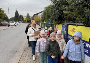 Grupa dzieci w jesiennych kurtkach i czapkach idzie parami chodnikiem z kostki wzdłuż ulicy pod opieką dorosłych, z których jedna niesie papierową torbę. Po prawej stronie, na ogrodzeniu porośniętym zielenią, wiszą żółto-granatowe banery reklamowe z napisem „AND BIURO”.