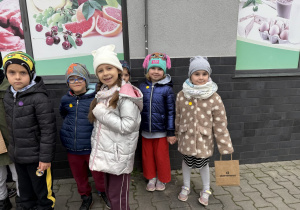Grupa dzieci stoi w rzędzie na chodniku z kostki brukowej przy ścianie budynku. Na ścianie wiszą plakaty z owocami, wędliną, jajkami i mlekiem, a jedno z dzieci trzyma papierową torbę z napisem „skierniewice”.