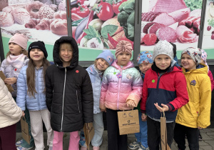 Grupa dzieci w kurtkach i czapkach pozuje do wspólnego zdjęcia na zewnątrz, przed ścianą z dużymi grafikami przedstawiającymi pieczywo, warzywa i wędliny. Kilkoro dzieci trzyma papierowe torby z napisem „skierniewice” i rysunkami owoców.
