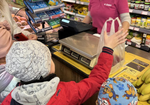 Dzieci przybijają piątkę ze sprzedawczynią w koszulce z napisem „U ŻAKA” przy kasie w sklepie spożywczym. Na ladzie stoi waga i leży foliowa reklamówka, a obok widać stojak z gumami Orbit, półki z produktami spożywczymi i banany.