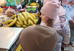 Grupa dzieci w czapkach i kurtkach stoi przy ladzie w małym sklepie spożywczym, na której leżą banany i waga sklepowa. W tle widać półki z pieczywem, przekąskami i innymi produktami z cenówkami, a jedno z dzieci trzyma papierową torbę z napisem „…erwice”.