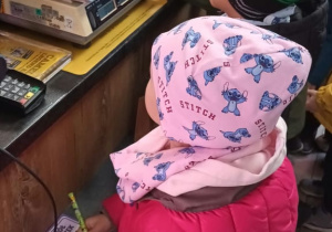 Dzieci w czapkach i kurtkach stoją przy ladzie w sklepie spożywczym. Jedno z nich zielonym pisakiem wypełnia kartkę z napisem „…ol Języczek”, a na ladzie stoją waga sklepowa i terminal płatniczy, za nimi widać banany i inne produkty.