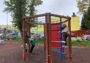 Dzieci bawią się przy drewnianym panelu edukacyjnym z liczydłem, kolorowymi koralikami na metalowych pętlach i obrotowymi klockami do gry w kółko i krzyżyk na placu zabaw z czerwoną nawierzchnią z gumowych płyt. W tle widać ogrodzenie z siatki, trawnik, drzewa, kosz na śmieci i boisko.