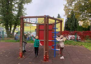 Dzieci bawią się na drewnianej konstrukcji do wspinaczki na placu zabaw z gumową nawierzchnią. Są tam drabinka, siatki linowe i niebieskie szczeble, a w tle rosną drzewa w jesiennych barwach i stoi żółty budynek.