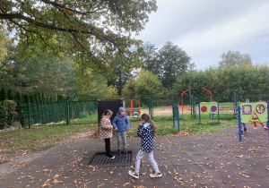 Dzieci bawią się na przedszkolnym placu zabaw o gumowej nawierzchni usianej jesiennymi liśćmi; dwoje stoi na trampolinie ziemnej, a w tle widać ogrodzenie, drzewa i tablice edukacyjne z napisem „PAPIER, KAMIEŃ, NOŻYCE”.