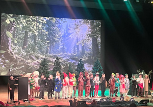 Dzieci w leśnych przebraniach i strojach bajkowych występują na scenie na tle grafiki z lasem
